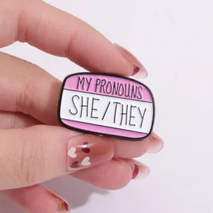 Pin’s pronoms She They tenu entre les doigts
