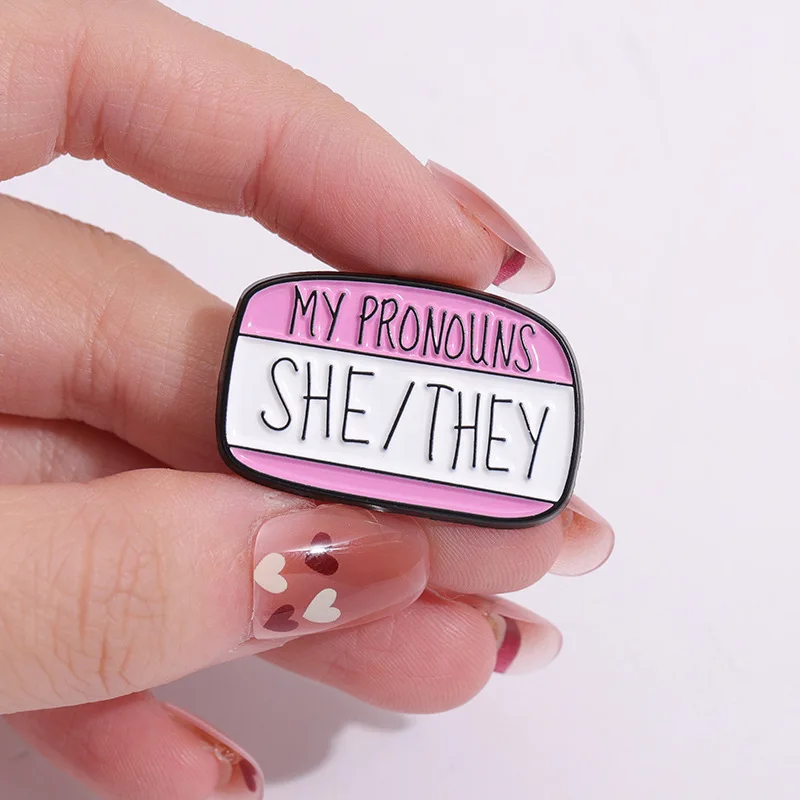 Pin’s pronoms She They tenu entre les doigts
